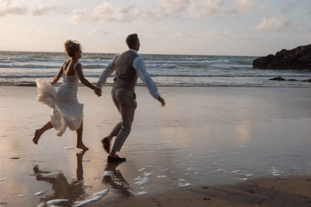 a married couple running along the beach. holding hands with a reflection of them on the sand. at Lusty wedding venue.