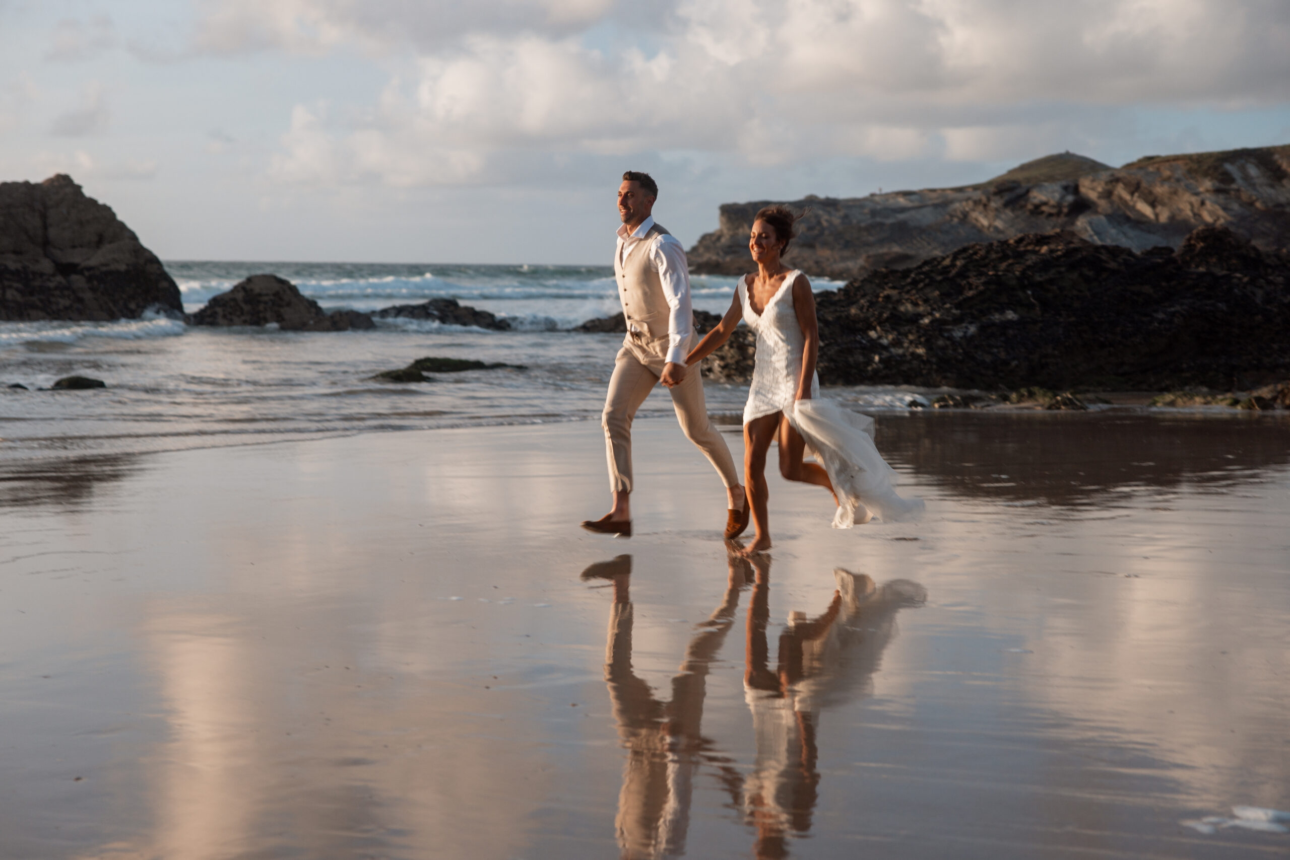 a married couple running along the beach. holding hands with a reflection of them on the sand. at Lusty wedding venue.